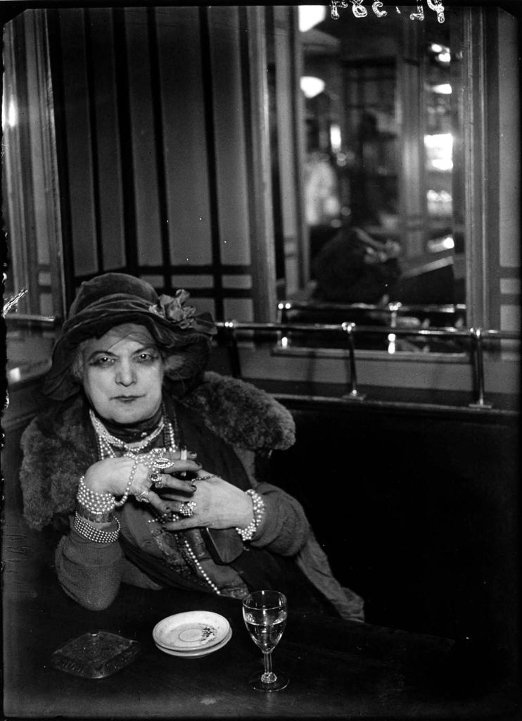 La Môme Bijou smoking a cigarette as she gazes into Brassaï's camera. She is wearing thick kohl eyeliner and masses of pearls around her neck and wrists. She is also wearing lots of rings.